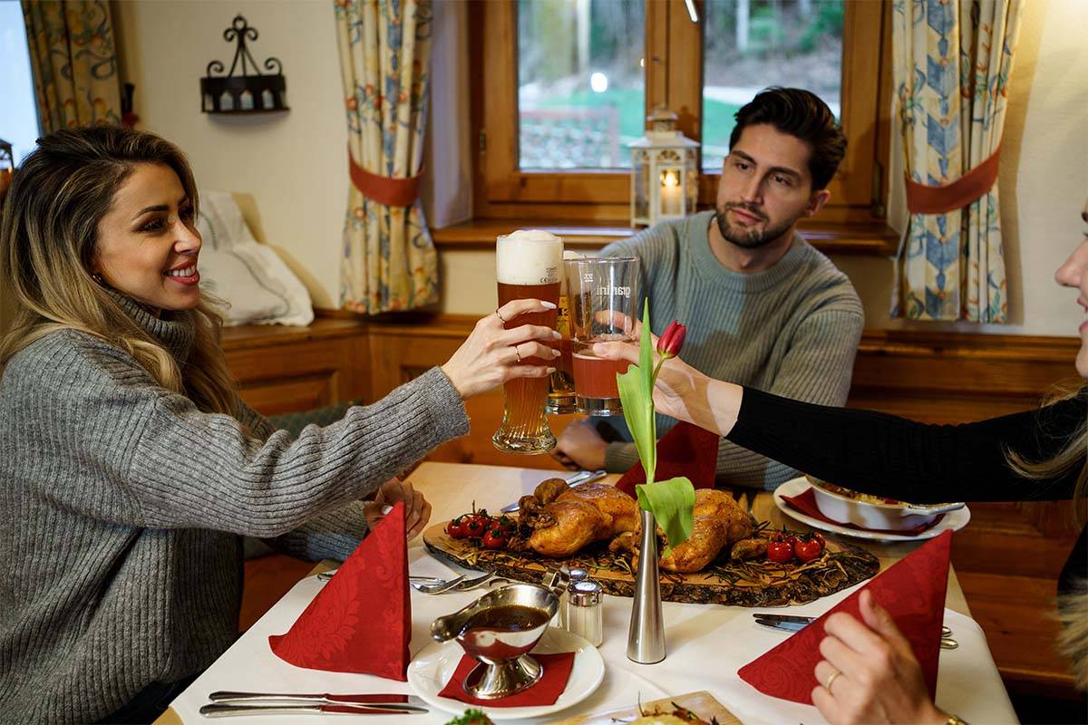 Drei Personen sitzen an einer Tischecke mit bayerischen Speisen und stoßen gemeinsam an.