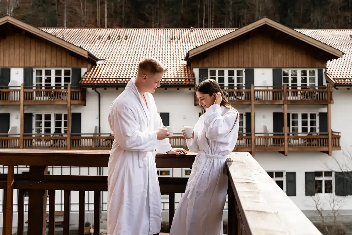 Ein Paar im Bademantel steht auf dem Balkon des Berghotel Hammersbach und genießt eine Tasse Kaffee.