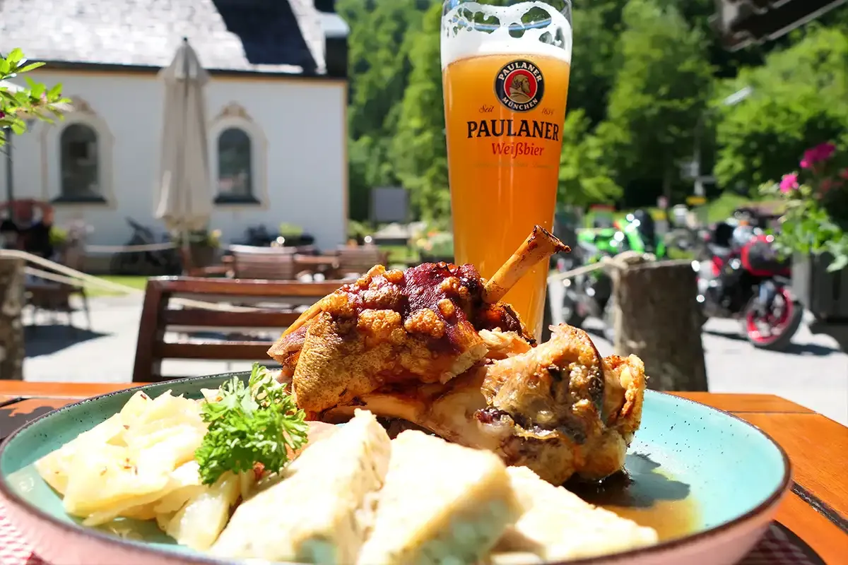 Schweinhaxe mit Beilage und Weißbier auf einem Tisch im Freien, umgeben von einer idyllischen Landschaft.