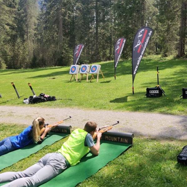 Zwei Personen liegen auf dem Boden im Grünen mit einem Biathlongewehr und schießen auf ein Ziel.