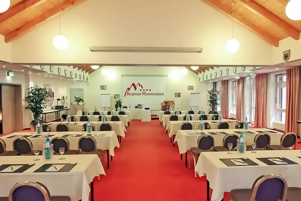 Großer Tagungsraum im Berghotel Hammersbach mit hohen Holzdecken, roten Teppischboden und Tischreihen.