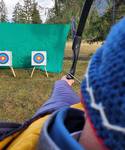 Eine Person zielt mit einem Bogen auf eine Zielscheibe im Freien. Im Hintergrund ist die Alpenlandschaft zu sehen.