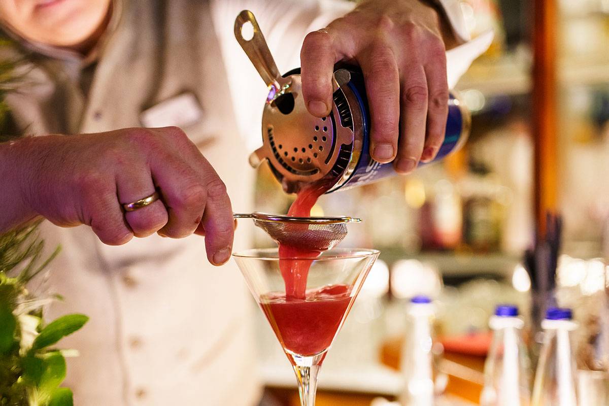 Ein Barmitarbeiter gießt den frisch geschüttelten Cocktail durch ein Sieb in das Cocktailglas.