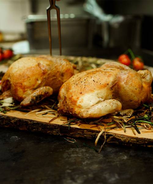 Zwei ganze Brathähnchen auf einer Holzplatte, umgeben von Gemüse, angerichtet.