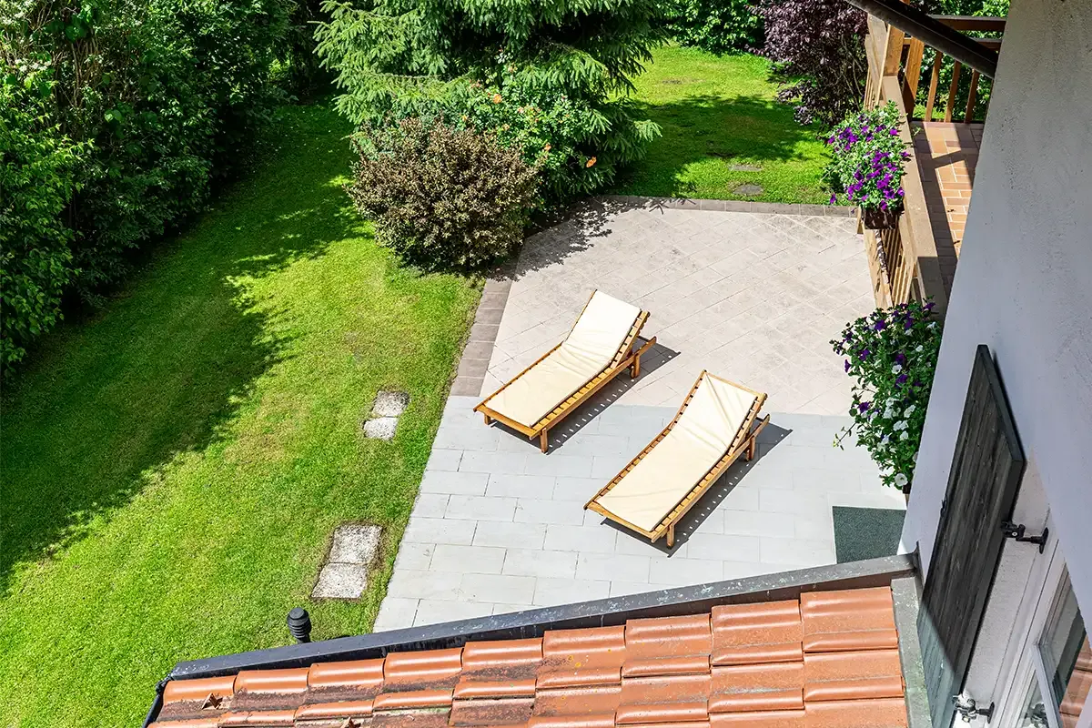 Aufnahme von oben auf die große, gepflasterte Terrasse am Chalet mit zwei Holz-Sonnenliegen umgeben vom Garten.