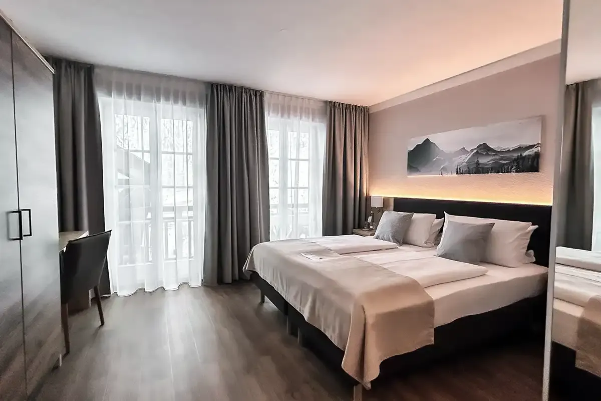 Großes und modernes Hotelzimmer mit Holzboden, Doppelbett, Fensterfront und geräumigem Kleiderschrank.