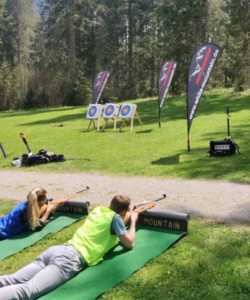 Zwei Personen liegen auf dem Boden im Grünen mit einem Biathlongewehr und schießen auf ein Ziel.