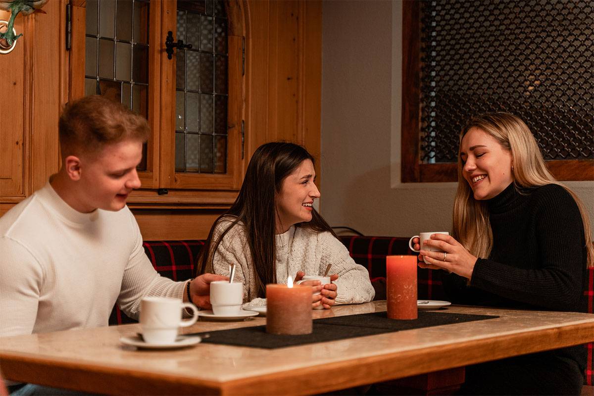 Drei Personen sitzen in einer Tischecke im Berghotel Hammersbach, genießen wärmende Heißgetränke und unterhalten sich.