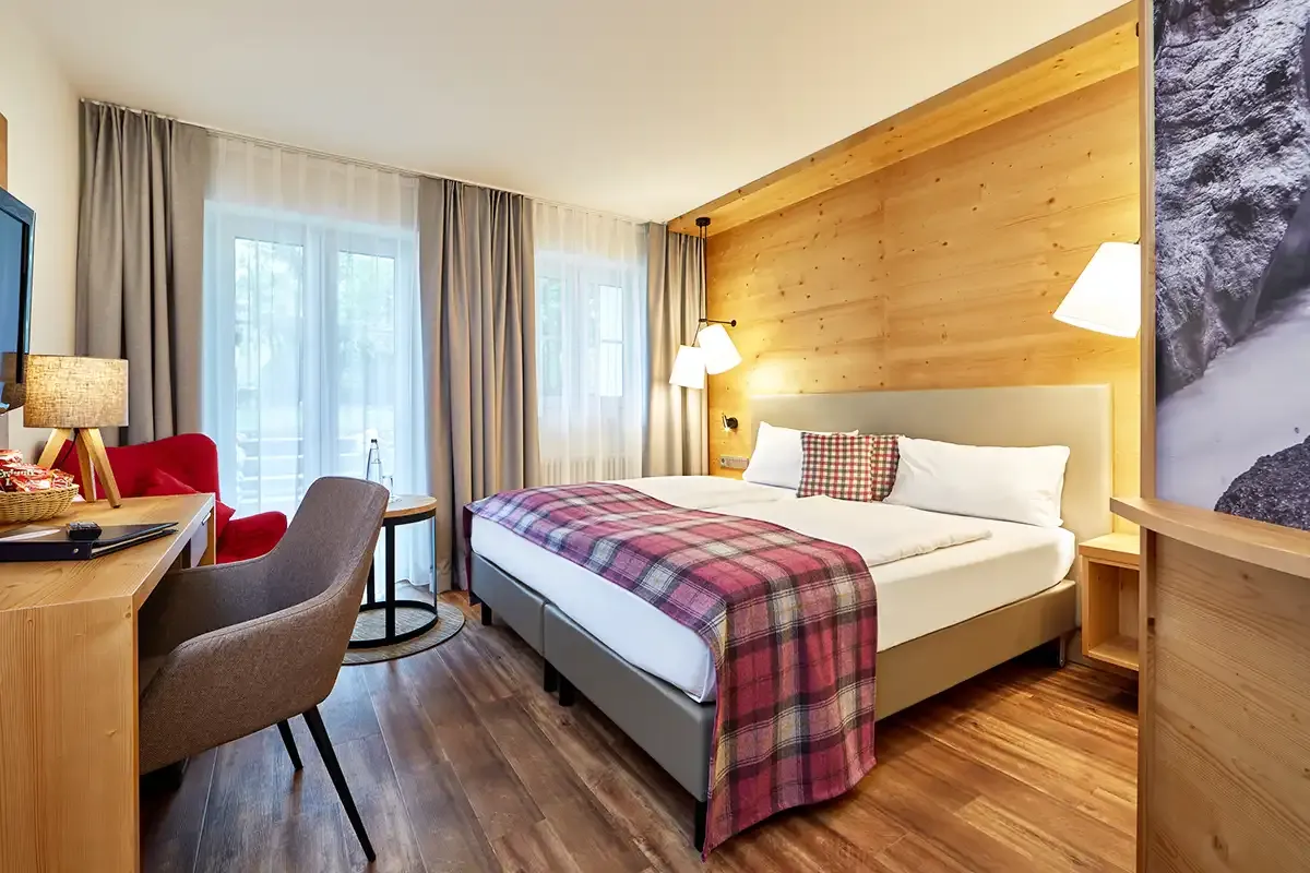 Gemütliches Doppelbett im Doppelzimmer mit moderner Ausstattung: Schreibtisch, TV, Sitzecke und Blick in die Berglandschaft.