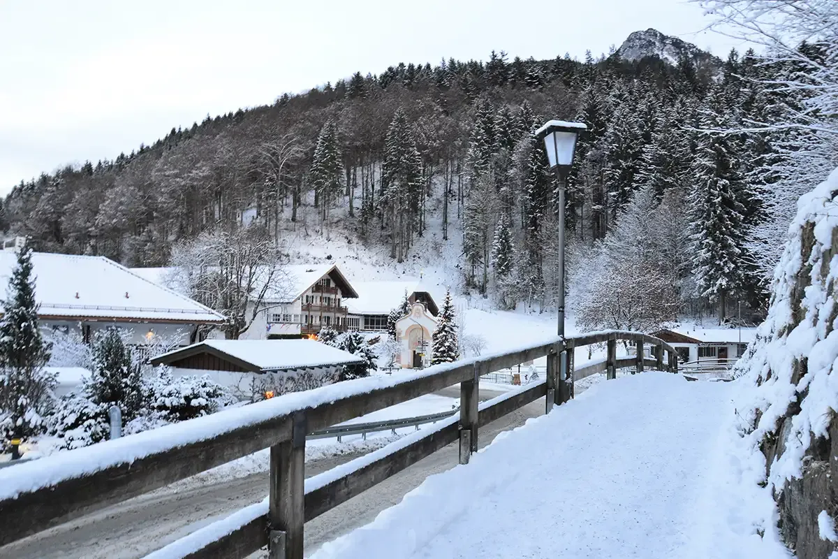 Verschneite Holzbrücke mit einer Ortschaft und Bergen im Hintergrund, umgeben von Tannen.
